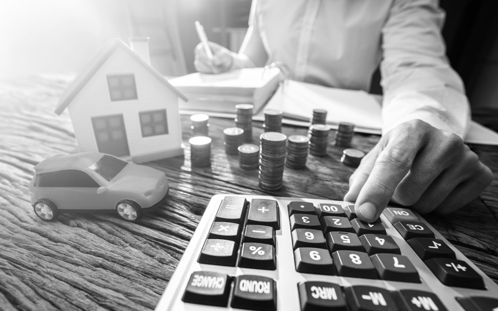 Person using a calculator with model house, toy car, and stacks of coins on a wooden desk, symbolizing rent calculator and property management.
