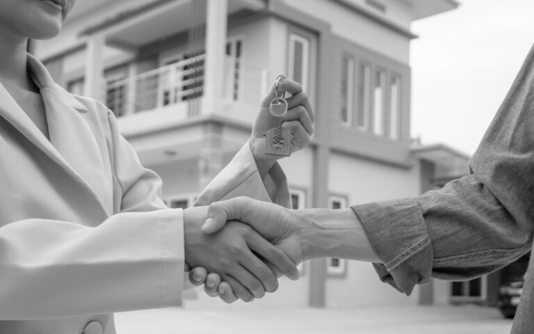 Close-up of a property manager and tenant shaking hands while holding a house key in front of a modern building.