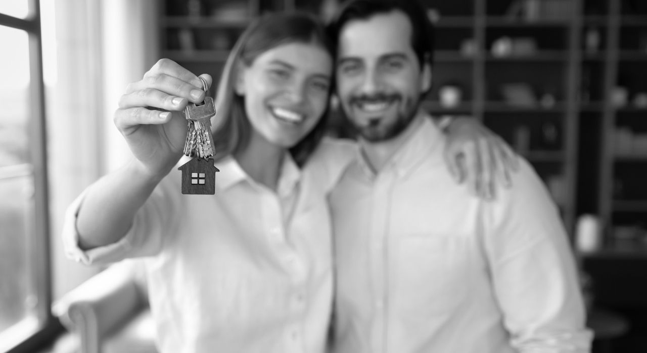 A smiling couple holds out a keychain shaped like a house, symbolizing a successful rental or new home.