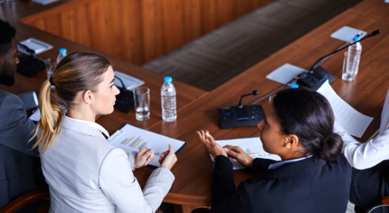 Two economists seated at a conference table reviewing printed charts and discussing economic forecasts during a formal meeting.