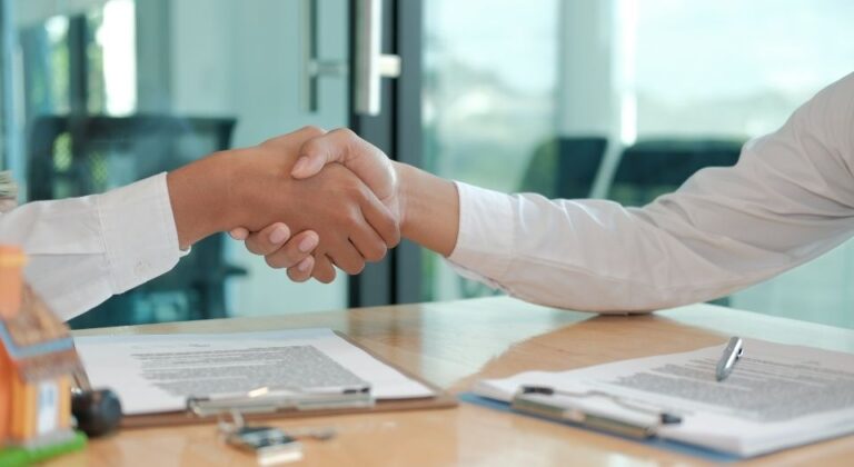 Two people shaking hands across a desk during a formal agreement, with documents, clipboards, and a small house model visible in the foreground.