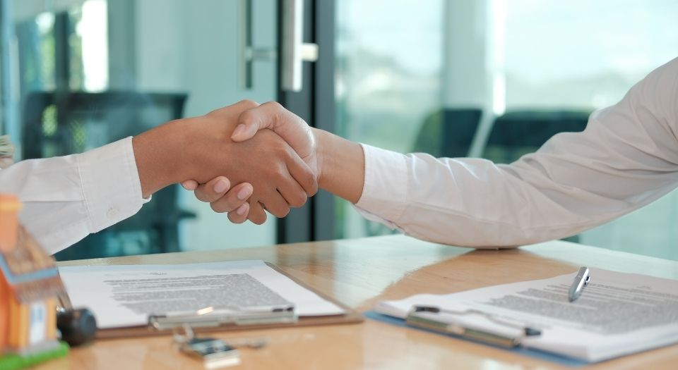 Two people shaking hands across a desk during a formal agreement, with documents, clipboards, and a small house model visible in the foreground.