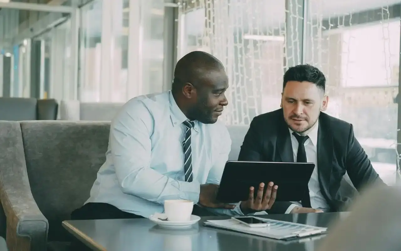 Two professional men in business attire sitting in a lounge area, collaborating and looking at a tablet together to discuss property strategy.
