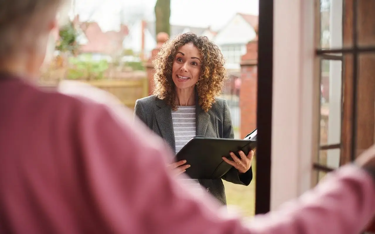 A friendly property manager holding a black folder and smiling while greeting a tenant or guest at the front door of a residential home.