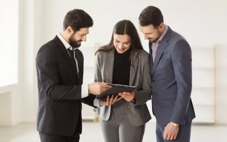 Three professionals in business attire, including a woman holding a clipboard, smiling and reviewing a rental agreement or lease document in a bright, modern office space.